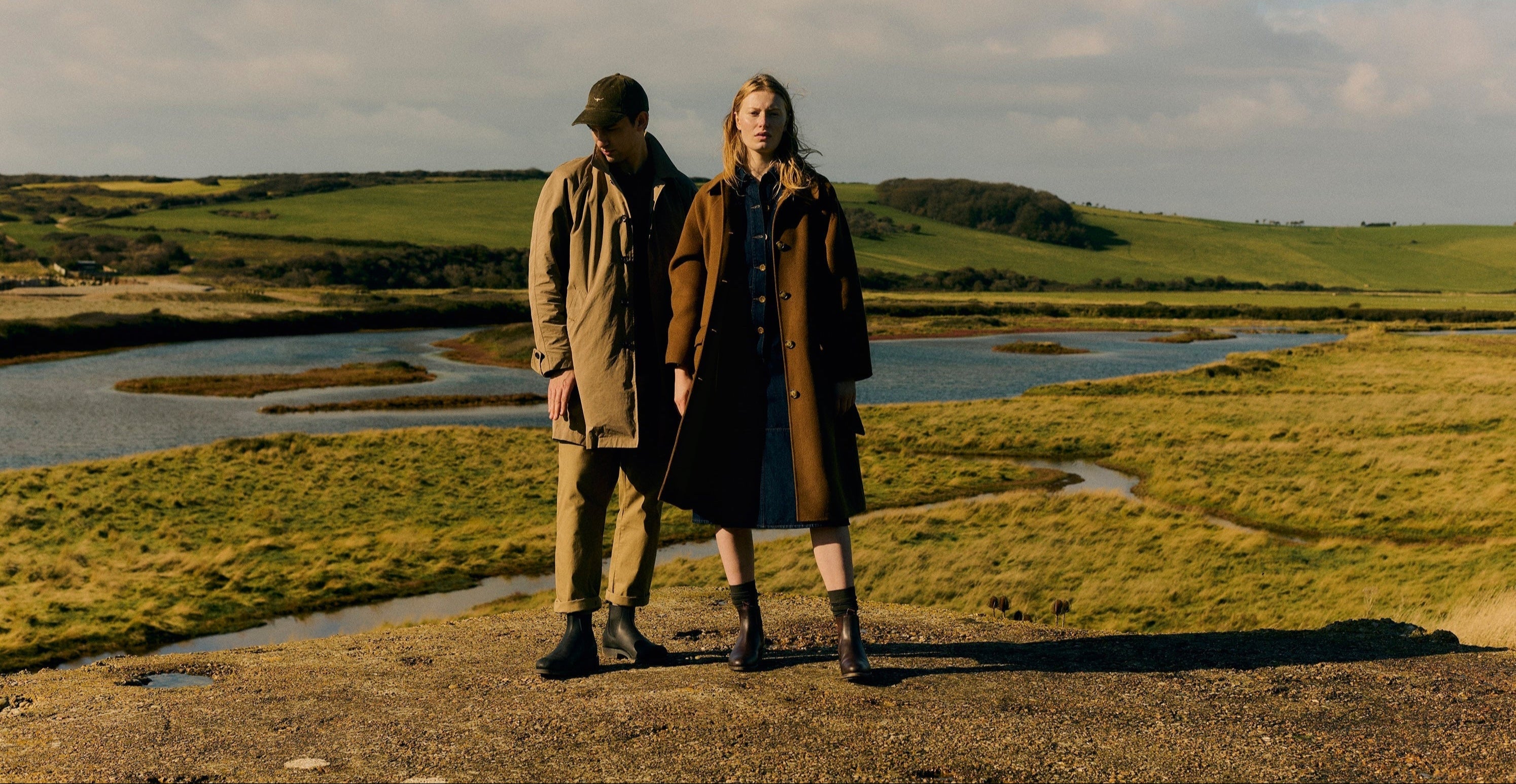 Two people standing on a grassy field with a river and hills in the background wearing RM Williams boots